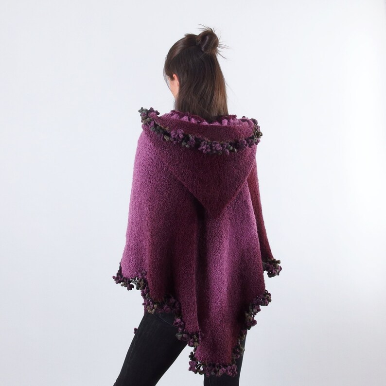 以下が含まれることがあります： 紫色のフード付きポンチョで、質感のあるニットとフリンジのトリムが付いています。フリンジは、紫と緑の色合いの小さなポンポンでできています。