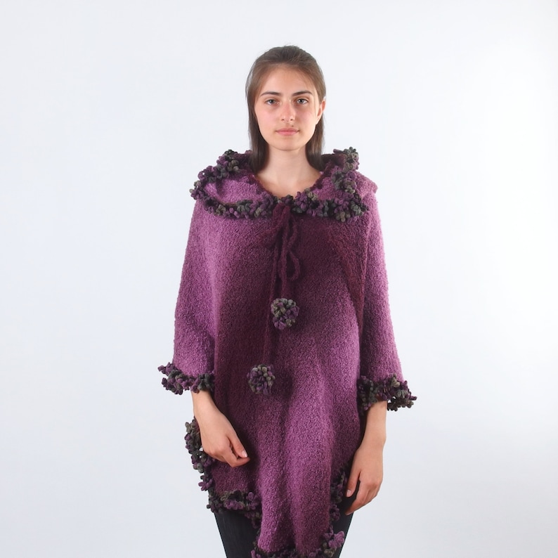 以下が含まれることがあります： ふわふわの質感と小さな紫色のポンポンで飾られたトリムが付いた、紫色のフード付きポンチョです。