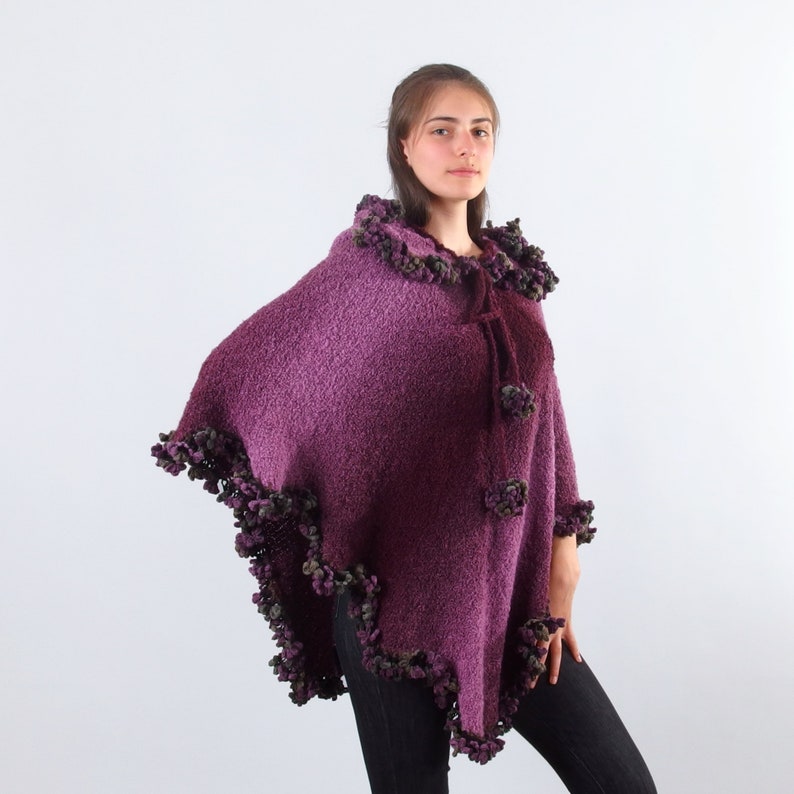 以下が含まれることがあります： フードと小さなポンポンの装飾的なトリムが付いた紫色のニットポンチョ。ポンチョは柔らかくふわふわした糸で作られており、濃い紫から薄い紫へのグラデーションになっています。