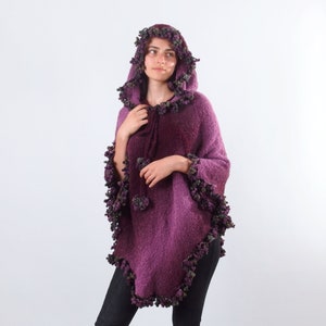 以下が含まれることがあります： パープルカラーのフード付きポンチョ。ふわふわの素材で、パープルとグリーンのポンポンが縁取りされています。