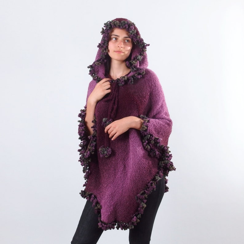 以下が含まれることがあります： パープルカラーのフード付きポンチョ。ふわふわの素材で、パープルとグリーンのポンポンが縁取りされています。