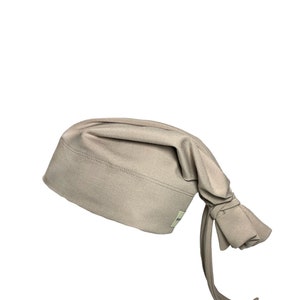 May include: A light brown surgical scrub cap with a long tie. The cap is made of a soft, stretchy fabric.