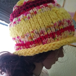 May include: A yellow, red, and white knitted hat with a fluffy texture. The hat has a wide brim and is made with a chunky yarn.