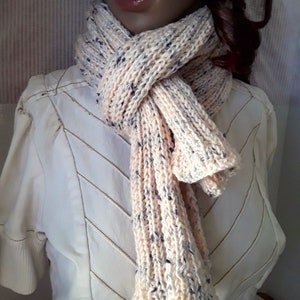 May include: A soft, knitted scarf in a cream color with peach and purple flecks. The scarf is tied around a mannequin's neck.