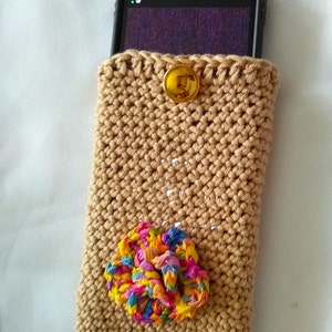May include: A crocheted phone case in a beige color with a gold button and a colorful fabric flower embellishment.