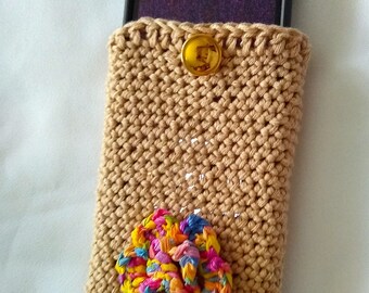 May include: A crocheted phone case in a beige color with a gold button and a colorful fabric flower embellishment.