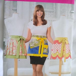 May include: A Simplicity sewing pattern for three different styles of aprons. The pattern is for one size and features a floral print, a blue and yellow print, and a red and white print.