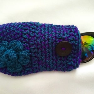 May include: Crocheted sunglasses case with a blue flower and a brown button. The case is made with purple and blue yarn.