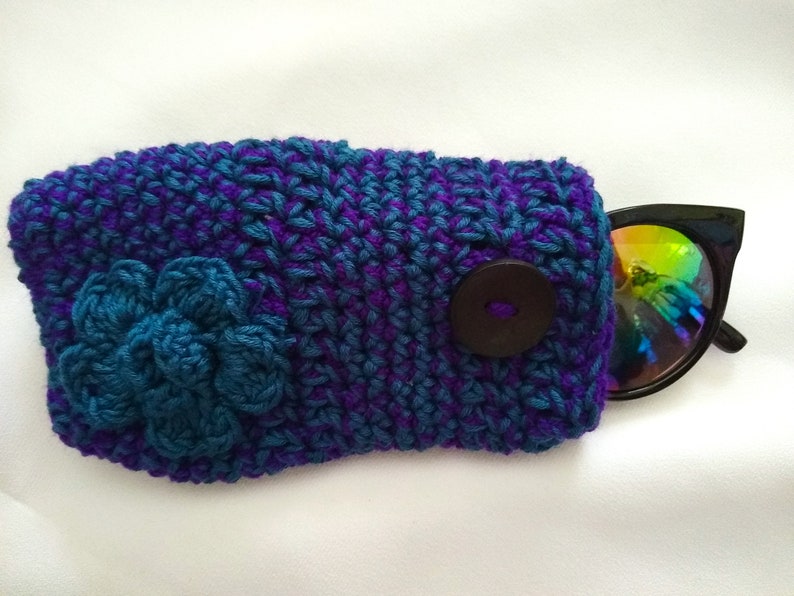 May include: Crocheted sunglasses case with a blue flower and a brown button. The case is made with purple and blue yarn.
