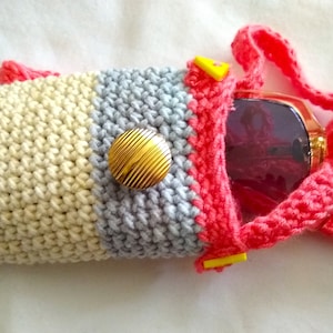 May include: A crocheted sunglasses case with a pink strap, a gold button, and a striped pattern in beige, cream, gray, and pink.