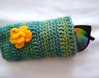 May include: A crocheted green and blue eyeglass case with a yellow flower embellishment. The case is holding a pair of sunglasses with iridescent lenses.