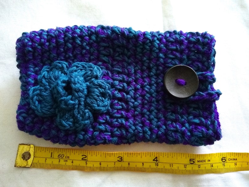 May include: A crocheted purple and teal phone case with a teal flower and a black button. The case measures 6 inches long.