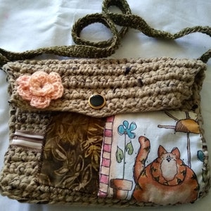 May include: Handmade crochet shoulder bag in shades of brown with a decorative peach-colored flower. The bag features a flap with a black button closure and a fabric panel with a cat illustration. The strap is a dark green.