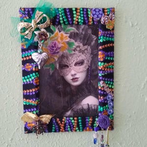 May include: A black picture frame with a purple, green, and orange beaded border. The frame has a picture of a woman wearing a masquerade mask. The frame is decorated with gold and silver embellishments.