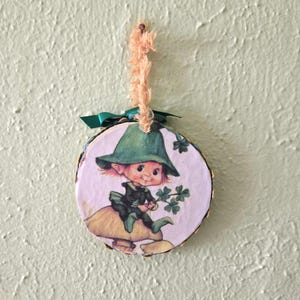 May include: A round wooden ornament featuring a cartoon leprechaun wearing a green hat and outfit, sitting on a mushroom and holding a sprig of shamrocks. The ornament has a gold-painted edge, a green ribbon, and a natural fiber loop for hanging.