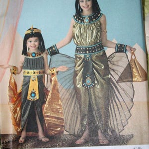 May include: Two girls wearing Egyptian-themed costumes. The costumes feature gold and black fabric, with turquoise and gold accents. The costumes include headbands, armbands, and flowing skirts. The background is a light blue color.
