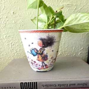 May include: A decorative ceramic flower pot featuring a whimsical illustration of a girl watering flowers. The pot has a white base with a colorful design, including bees and floral accents. The pot holds a green plant.