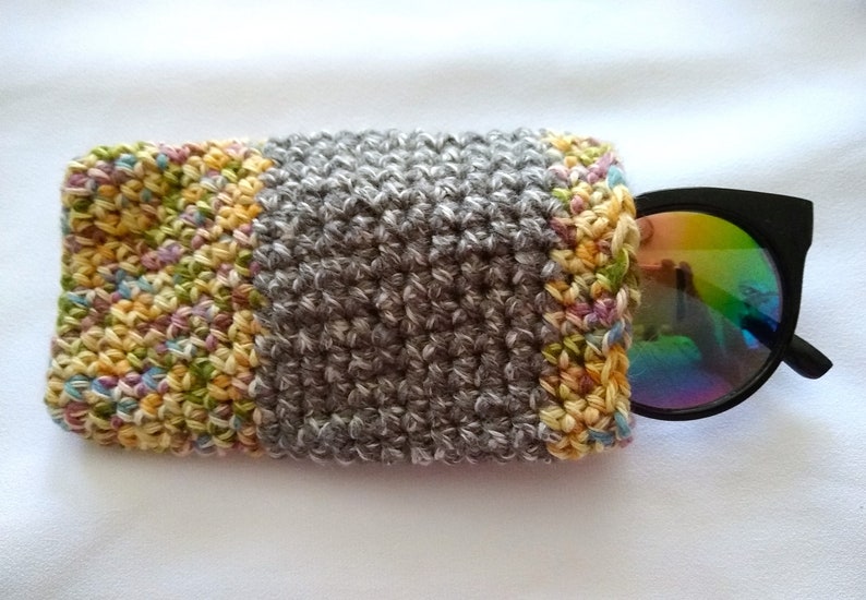May include: A gray crocheted eyeglass case with a colorful crocheted border. The case is open and a pair of black sunglasses with iridescent lenses are partially inside.