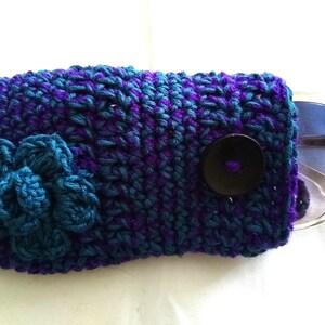May include: Crocheted sunglasses case with a blue flower and a black button. The case is made with purple and blue yarn.