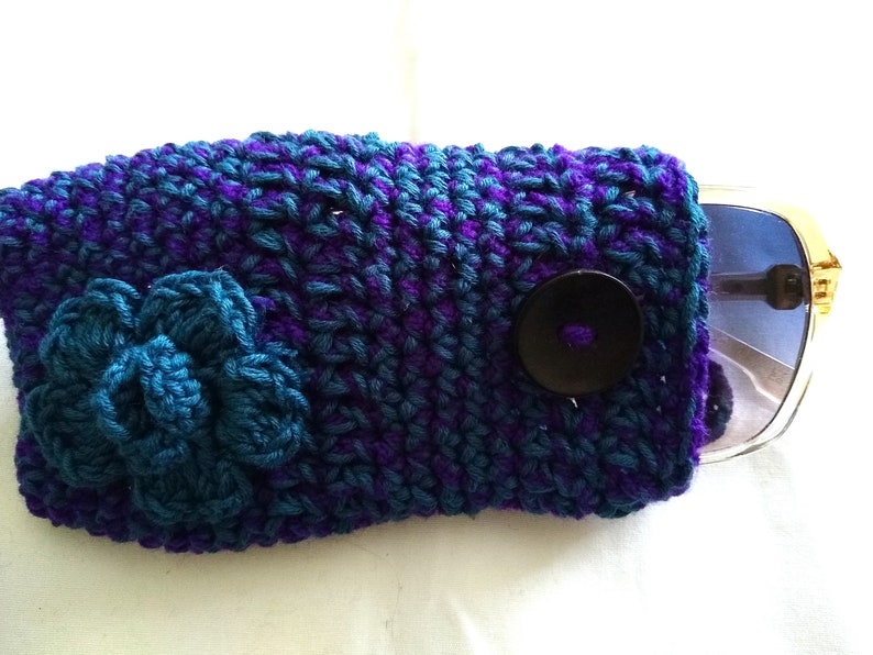 May include: Crocheted sunglasses case with a blue flower and a black button. The case is made with purple and blue yarn.