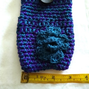 May include: A crocheted phone case in shades of purple and teal with a teal crocheted flower on the front. The case is measured with a ruler, showing it is approximately 2 inches wide.