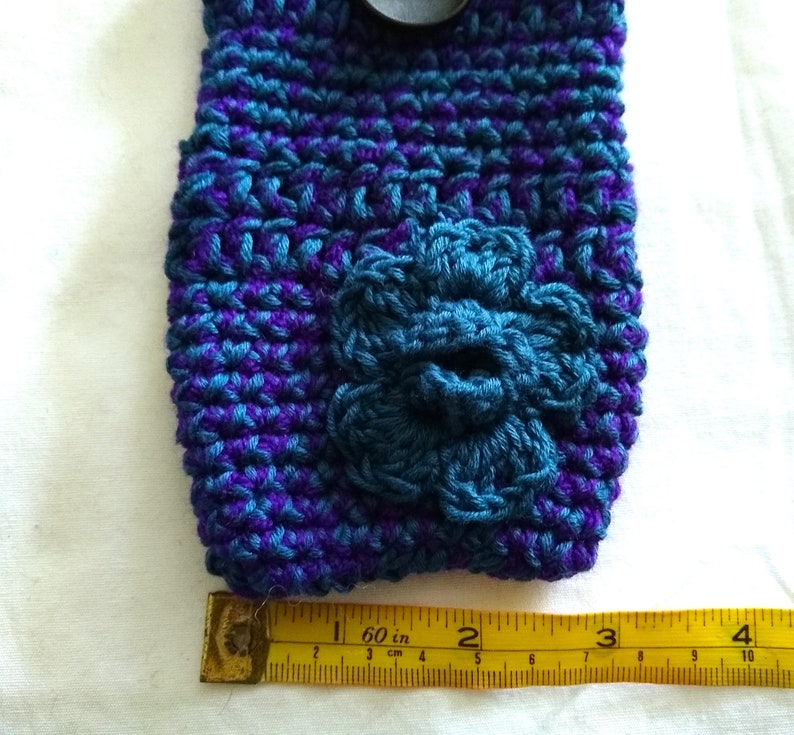 May include: A crocheted phone case in shades of purple and teal with a teal crocheted flower on the front. The case is measured with a ruler, showing it is approximately 2 inches wide.