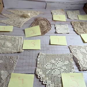 May include: A collection of vintage crochet doilies and lace pieces in various shapes and patterns. The pieces are in shades of cream and beige, arranged on a light wooden surface. Each piece is labeled with a number on a yellow sticky note.