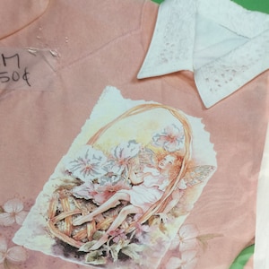 May include: A pink sweatshirt with a white collar and a printed image of a fairy sitting in a basket of flowers. The text "M 50$" is visible on the sweatshirt.