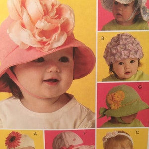May include: A collection of baby hats in various styles. One pink hat features a large peach-colored flower. Other hats include a white hat with a red flower, a lavender floral hat, a green hat with yellow flowers, and a white headband with a flower.