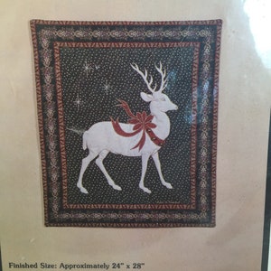 May include: A square Christmas-themed quilt panel featuring a white reindeer with a red bow, set against a dark background with stars. The border is a detailed pattern in red and black. Finished size is approximately 24" x 28".