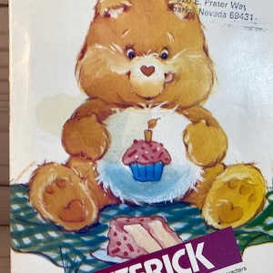 May include: A vintage Care Bear birthday pattern featuring a brown bear holding a pink and blue cupcake. The bear is sitting on a green and white checkered picnic blanket with a slice of cake in front of it. The pattern is titled "Butterick" and includes the copyright information "MCMLXXXIII American Greetings Corporation".