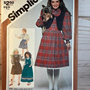 May include: Vintage Simplicity sewing pattern for girls' dresses, size 7 & 8. The cover features a girl in a red plaid dress with a black vest, holding a small dog. Additional dress designs are shown in the lower left corner. The pattern includes a special chart on how to work with plaids.