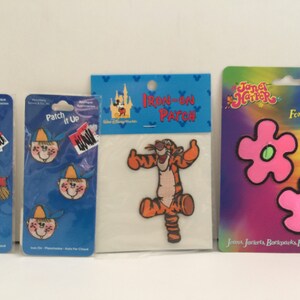 May include: Three packages of iron-on patches. The first package has a blue background with a cartoon boy wearing a baseball cap. The second package has a blue background with a cartoon tiger. The third package has a purple background with two pink flower-shaped patches.