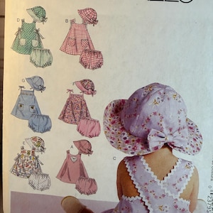 May include: A McCall's sewing pattern featuring various baby and toddler outfits, including dresses, bloomers, and sun hats. The outfits are in pastel colors and floral patterns. The pattern includes the text "McCall's" at the top.