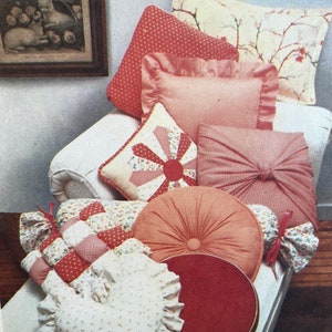 May include: A collection of decorative pillows in various shapes and patterns. Pillows include a heart-shaped pillow with ruffled edges, a round pillow with a button center, and pillows with floral and polka dot designs. The pillows are in shades of pink, red, and cream.