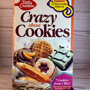 May include: A vintage Betty Crocker cookbook titled "Crazy About Cookies" featuring images of various cookies. The cover includes text that reads "Exclusive! 12 Fun Cookie Patterns" and "7 Cookies from 1 Mix!" The cookbook is displayed on a black stand.