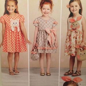 May include: A sewing pattern envelope for Ruby Jean's Closet featuring three dress variations with animal motifs, including a cat-themed orange floral dress, a grey lattice print dress with a large bow, and a floral dress with bunny ear accents.