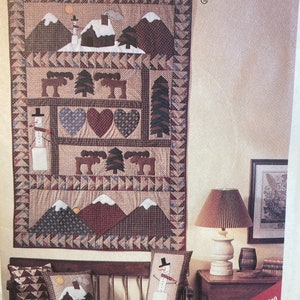 May include: A quilted wall hanging with a winter scene, featuring mountains, a house, and a snowman. The design includes hearts, trees, and moose. The text "AMERICAN TRADITION" is at the top. Two matching pillows with winter scenes are on a bench.