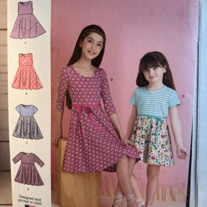 May include: A Simplicity S9322 sewing pattern envelope for children's dresses in sizes 3-6. The cover features illustrations of four dress variations and a photograph of two children wearing a pink patterned dress and a striped floral dress.