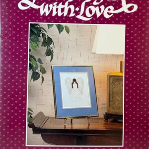 May include: A book cover titled Hardanger with Love by Janice Love featuring a dark magenta background with a repeating pattern of small pink hearts. The center displays a framed needlework piece of an angel with brown hair and white wings.