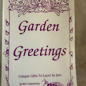 May include: A white book cover titled Garden Greetings by Jackie Gannaway with the subtitle Unique Gifts To Layer In Jars. The cover features purple line art of a jar lid, garden tools, ladybugs, and a bee, all in a purple ink design.