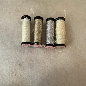 May include: Four spools of metallic embroidery thread arranged in a row. The spools feature thread in shades of pale cream, antique gold, bright silver, and light champagne. Each spool has a black plastic base with a small pink circular label.