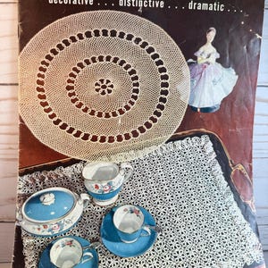 May include: Vintage book cover titled "Doilies" with the words "decorative, distinctive, dramatic." The cover features a crocheted doily, a porcelain figurine, and a tea set with blue and floral details. The tea set includes a covered dish, creamer, and two teacups with saucers.