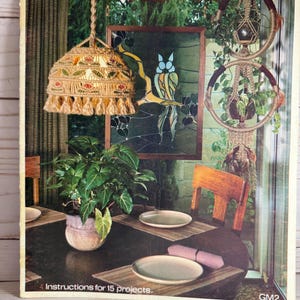 May include: A craft book cover titled MACRAME with a PURPOSE featuring a macrame lampshade with red and green floral patterns, a hanging macrame plant holder with two circular rings, a potted green plant, and two plates on a dining table.