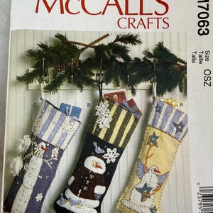 May include: McCall's Crafts M7063 sewing pattern envelope featuring three decorative Christmas stockings. The stockings display snowman and star motifs in blue, black, and cream colors, hanging from a green pine garland.