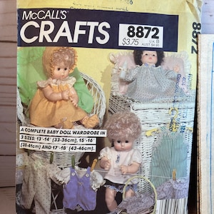 May include: A vintage McCall's Crafts sewing pattern for a baby doll wardrobe. The pattern includes instructions for making a variety of outfits for dolls measuring 13-14 inches (33-36 cm), 15-16 inches (38-41 cm), and 17-18 inches (43-46 cm). The pattern cover features illustrations of dolls wearing the different outfits.