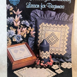 May include: A Designs by Gloria & Pat instructional leaflet titled A Hardanger Embroidery Lesson for Beginners. The cover features a dark blue background with a floral arrangement, a wooden box, a dark blue candle, and Hardanger embroidery.