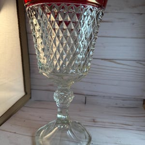 May include: A clear glass goblet with a red rim. The goblet has a diamond pattern on the bowl and a decorative stem. The base is wide and rounded. The goblet is likely vintage and suitable for serving beverages.