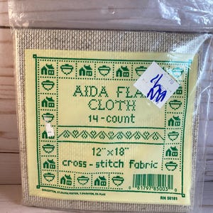 May include: Aida cloth for cross-stitch, 14-count, 12 inches by 18 inches. The fabric is beige and has a green border with a pattern of houses and bowls. The label says "Aida Flax Cloth 14-count" and "cross-stitch Fabric".
