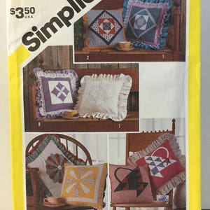 May include: Simplicity sewing pattern number 6146 for making four different pillow designs. The pillows are shown on a wooden bench and a rocking chair. The pillows are made with a variety of fabrics and colors, including red, blue, yellow, green, and white. The pattern includes instructions for making the pillows and the ruffles.